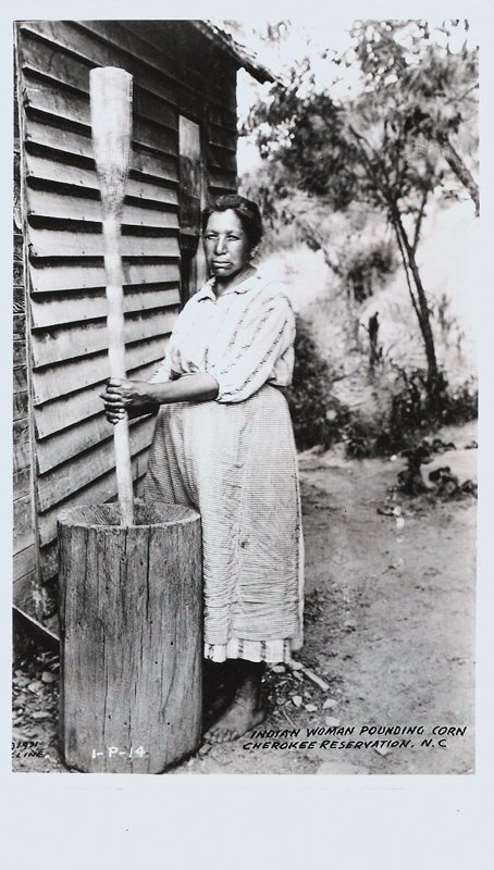 A Cherokee woman uses a large wooden pestle and mortar to pound corn into grain.