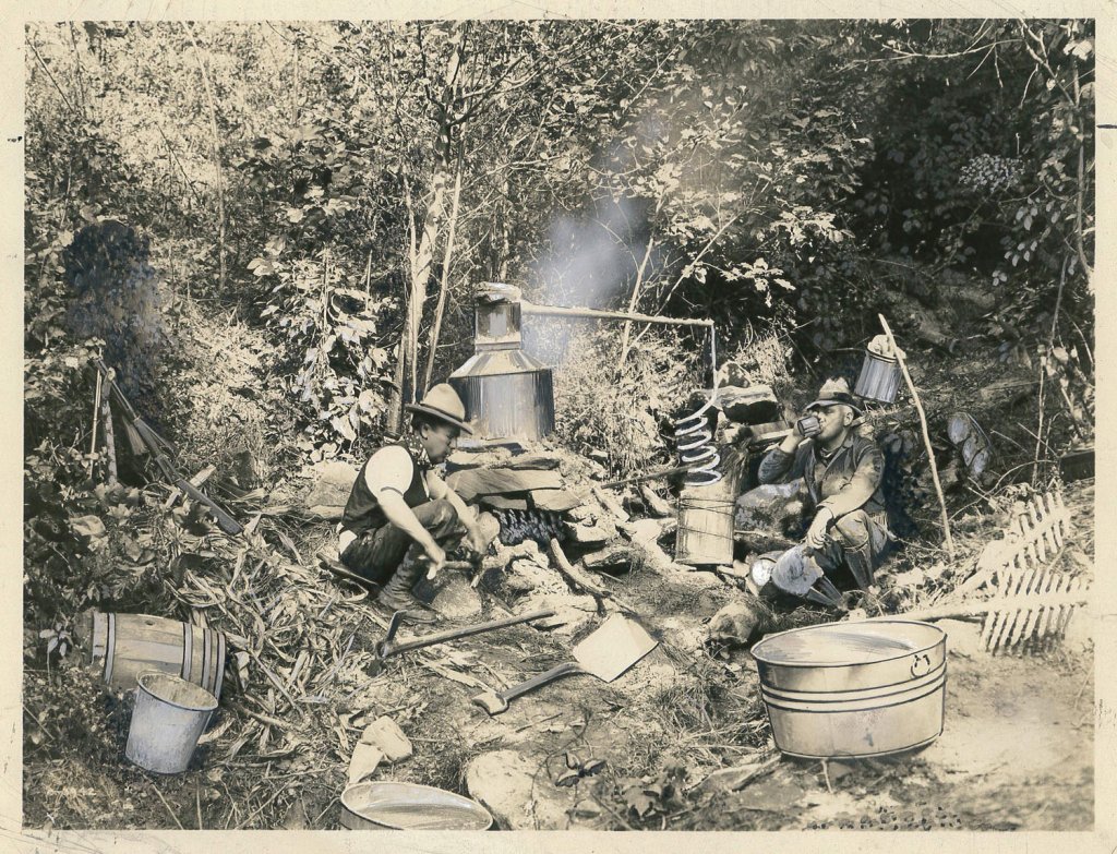 A souvenir postcard shot by George Masa shows a staged moonshine setup. Historic figure Bascom Lamar Lunsford sits on the left. Lunsford was a lawyer and Appalachian folk musician.