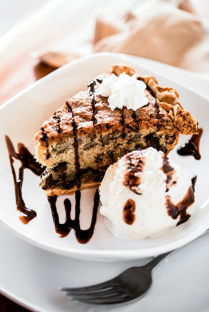 A slice of deep-dish chocolate chip pie with pecans, served warm, is accompanied by a scoop of vanilla ice cream.