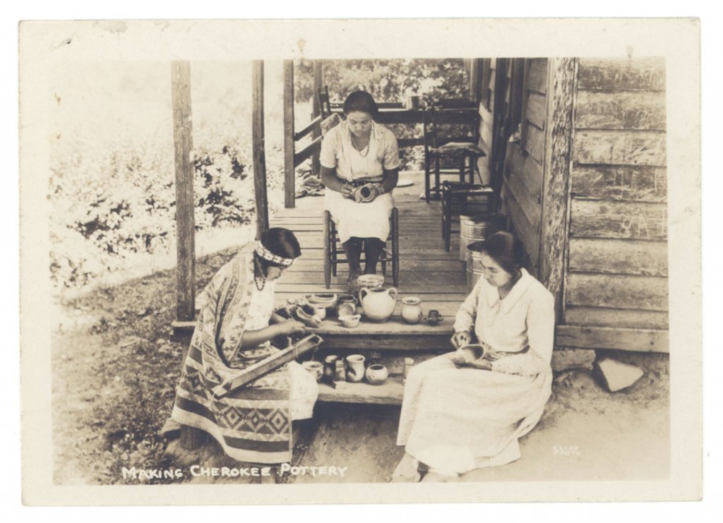 Apostcard of three Cherokee women crafting on the porch. Two are burnishing pottery, and one is using a handloom.
